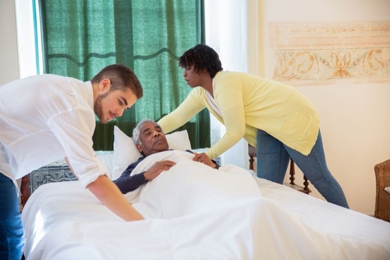 Two carers attending to an older man resting in bed at home